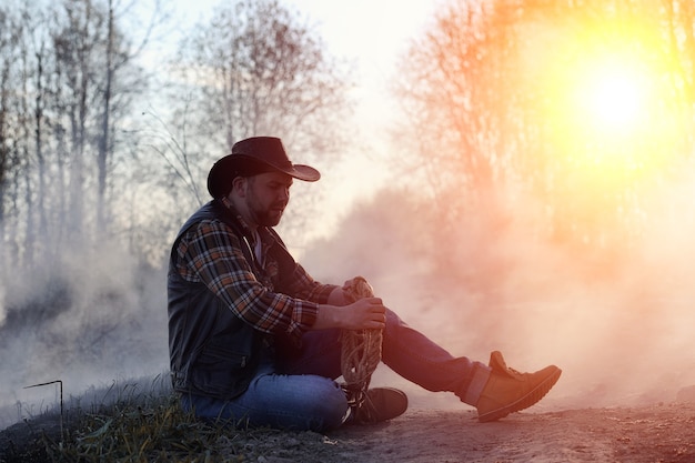 Un uomo indossa un cappello da cowboy e un loso nel campo. Contadino americano in un campo che indossa un cappello di jeans e con un lazo nel fumo di un incendio. Un uomo cammina in un campo in fiamme con un cappello