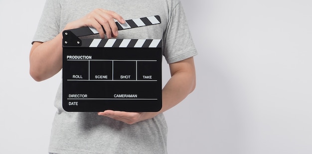 Un uomo indossa una maglietta grigia e la mano tiene in mano un batacchio nero o una lavagna per film. utilizza nella produzione video o nell'industria cinematografica. È uno sfondo bianco.