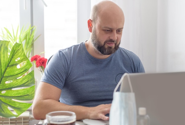 Un uomo lavora a casa con un laptop, una maschera protettiva per il caffè e una quarantena disinfettante a causa della pandemia di coronavirus
