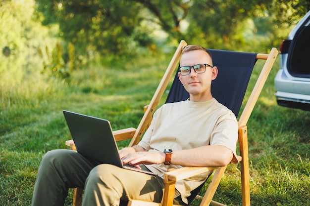 Un uomo libero professionista si siede su una sedia di legno nella natura e lavora online su un laptop Un uomo viaggia e lavora in remoto su un computer portatile Lavoro d'ufficio nella natura Vacanze