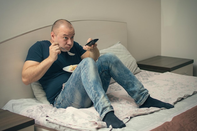 Un uomo mangia a letto e tiene in mano il telecomando della TV