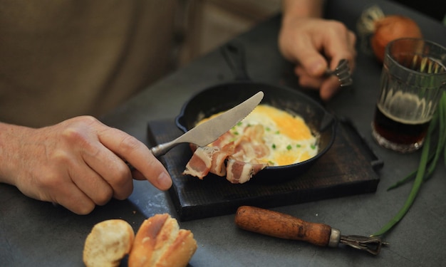 Un uomo mangia uova e pancetta da una padella di ghisa e beve birra scura