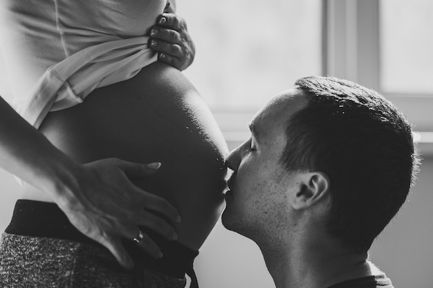 Un uomo, marito, bacia la pancia della moglie incinta vicino alla finestra di casa. Gravidanza familiare felice, aspettativa. Foto in bianco e nero.