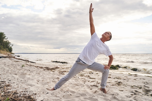 Un uomo maturo calmo si sta allenando al mattino sulla riva del mare e pratica asana con la mano alzata verso il cielo sky