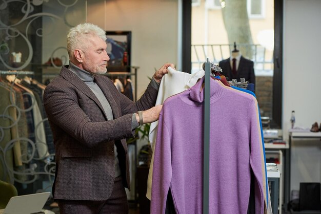 Un uomo maturo con i capelli grigi sceglie un maglione a collo alto in un negozio di abbigliamento Un cliente maschio in un abito di lana in una boutique