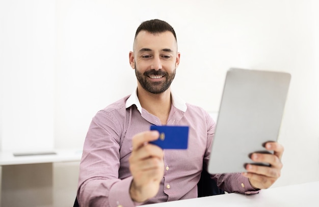 Un uomo maturo e felice in camicia si siede al tavolo con un tablet e usa una carta di credito.