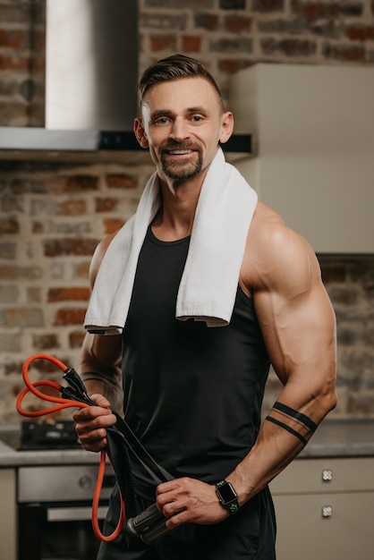Un uomo muscoloso felice con un asciugamano bianco sulle spalle posa con la corda elastica nel suo appartamento
