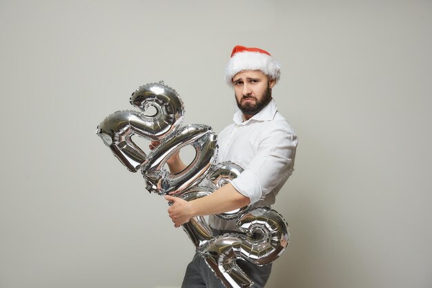 Un uomo orgoglioso con un cappello da Babbo Natale di velluto rosso tiene in mano palloncini d'argento a forma di 2022. Un ragazzo felice con la barba a una festa di Capodanno.