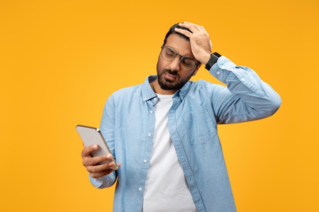 Un uomo perplesso con una camicia blu tiene in mano il suo smartphone e lo guarda con un'espressione confusa