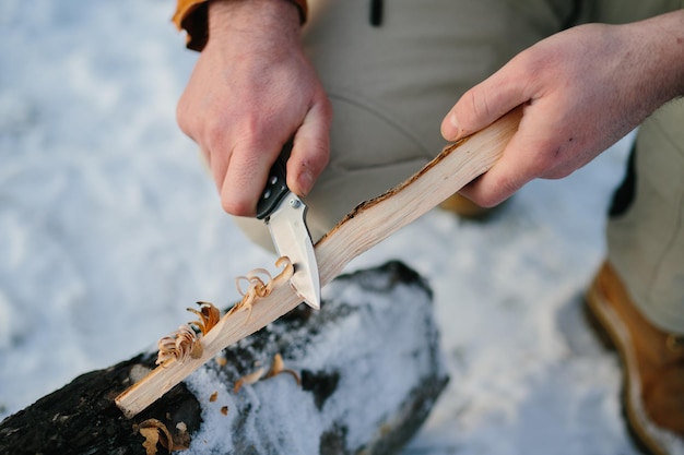 Un uomo prepara la lana di legno con un coltello per accendere un fuoco in una foresta invernale al tramonto Concetto di sopravvivenza invernale