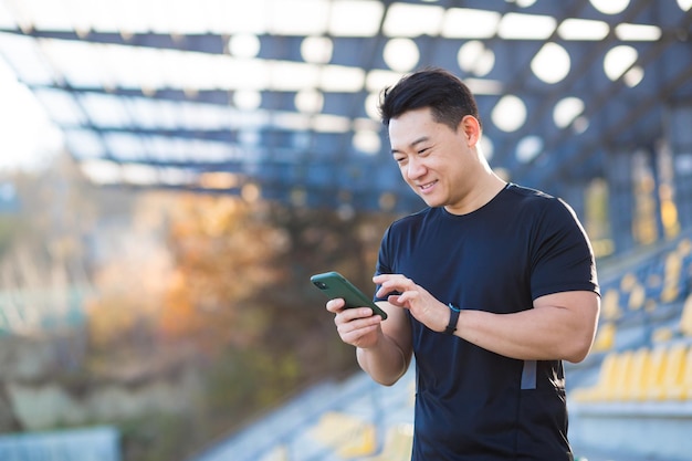 Un uomo sano guarda un video su un telefono cellulare dopo un allenamento mentre è seduto all'aperto utilizzando un'app su un dispositivo wireless 4g L'atleta asiatico è felice e sorridente dopo aver corso a riposo
