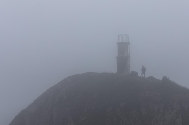 Un uomo scala la montagna fino a un faro abbandonato in un pesante fogman in cima a una montagna in un faro nella nebbia