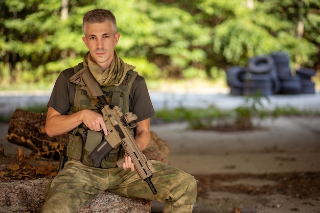 Un uomo seduto con un fucile d'assalto softair in uniforme militare in un hangar di cemento