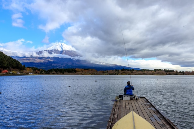 Un uomo seduto e pesca da solo sul ponte di legno al lago Kawaguchiko in Giappone durante il tramonto con Fuji Mountain Background