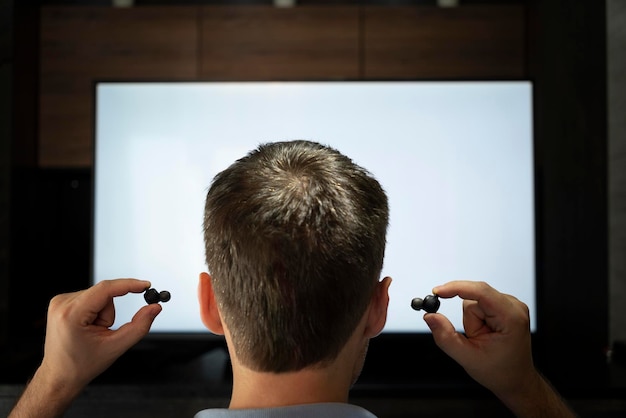 Un uomo si infila le cuffie nell'orecchio sullo sfondo di uno schermo televisivo che guarda la TV con le cuffie di notte