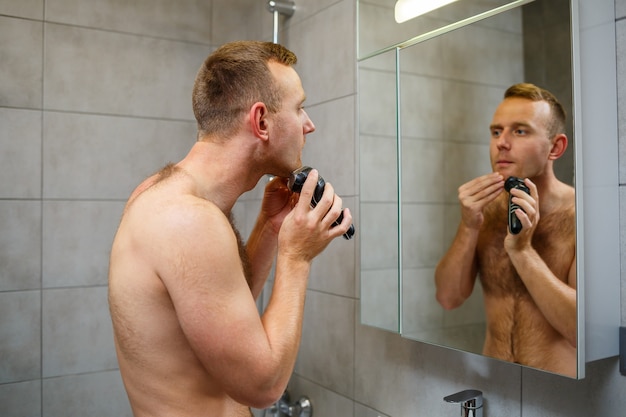 Un uomo si rade la faccia con un rasoio elettrico davanti a uno specchio. Irritazione della pelle. Procedura del bagno