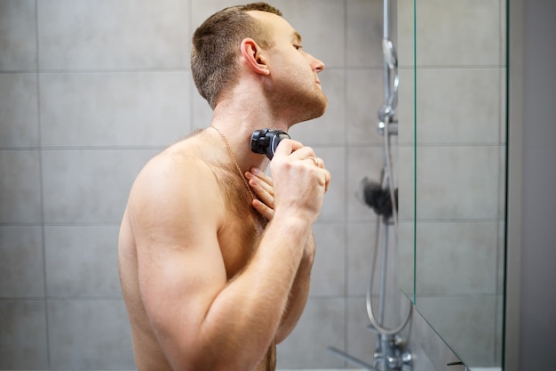 Un uomo si rade la faccia con un rasoio elettrico davanti a uno specchio. Irritazione della pelle. Procedura del bagno