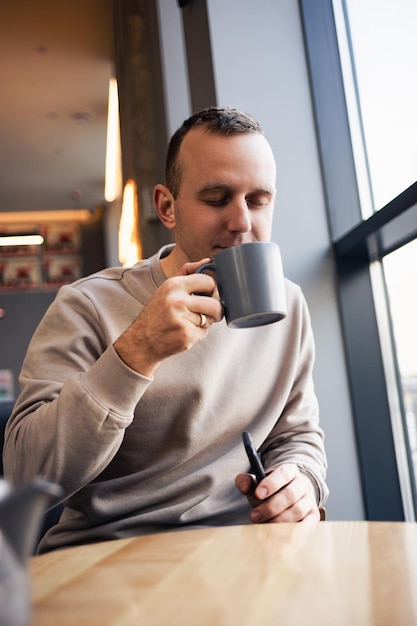 Un uomo solitario e attraente si siede al tavolo di un bar, beve un tè caldo profumato. Ragazzo sorridente in abiti casual si siede in un bar e beve caffè