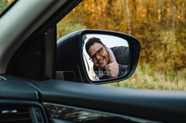 Un uomo sorridente nello specchietto laterale dell'auto mostra un grosso bastone dietro la foresta autunnale