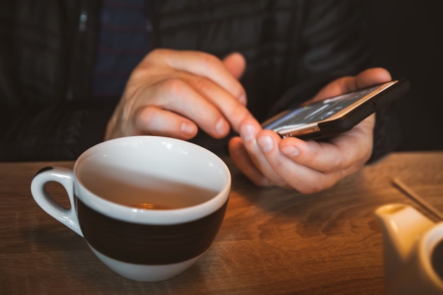 Un uomo sta riposando in un bar, bevendo tè, leggendo e scrivendo messaggi su uno smartphone. Lavoro in ufficio mobile. Comunicazione per tutto il giorno Ñ. Immagine ravvicinata con messa a fuoco morbida e profondità di campo ridotta