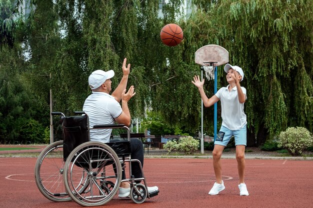 Un uomo su una sedia a rotelle gioca a basket con suo figlio sul campo sportivo. Genitore disabile, infanzia felice, concetto di persona disabile.