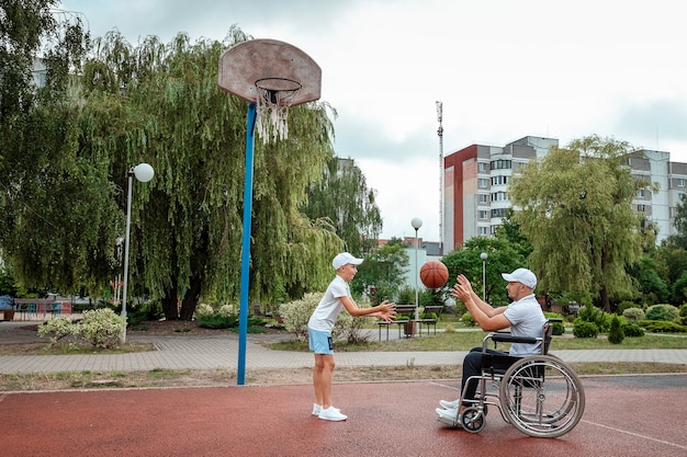 Un uomo su una sedia a rotelle gioca a basket con suo figlio sul campo sportivo. Genitore disabile, infanzia felice, concetto di persona disabile.