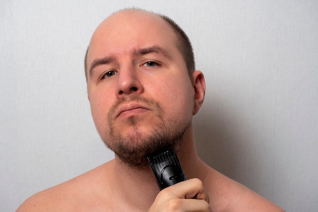 Un uomo su uno sfondo grigio si rade la barba con un rasoio elettrico. Guarda la telecamera, tagliandosi i capelli. La bellezza e la cura degli uomini a casa.