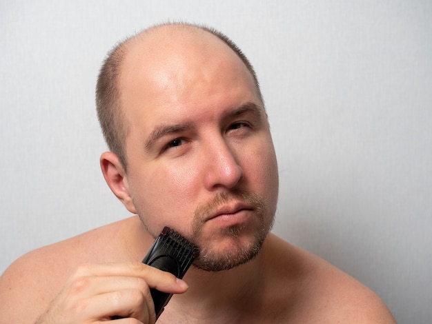 Un uomo su uno sfondo grigio si rade la barba con un rasoio elettrico. Guarda la telecamera, tagliandosi i capelli. La bellezza e la cura degli uomini a casa.