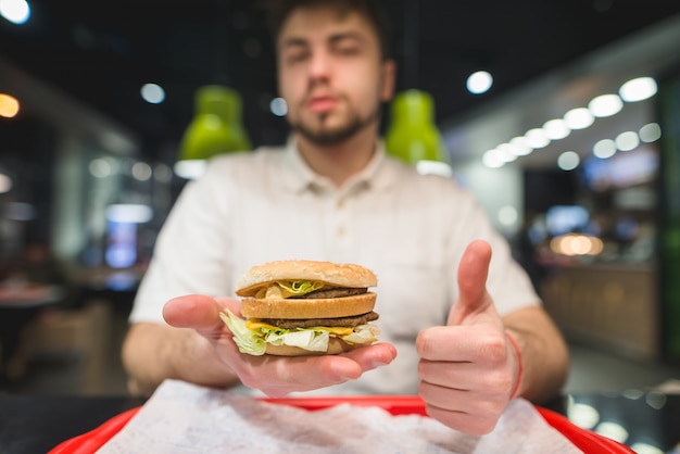 Un uomo tiene in mano un grosso hamburger e mostra il pollice in su. A un uomo piace il fast food. Un gustoso hamburger nelle tue mani.