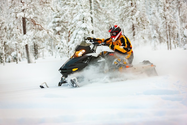 Un uomo travestito da corridore con una tuta giallo-nera e un casco rosso-nero, alla guida di una motoslitta ad alta velocità attraverso la neve profonda sullo sfondo di una foresta innevata.