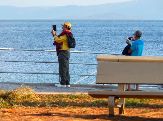 Un uomo vestito alla moda con un cappello scatta una foto del mare sull'argine in Grecia