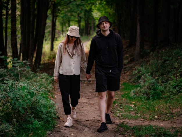 Un uomo vestito di nero e una donna con una camicia beige e un cappello camminano tenendosi per mano nella foresta