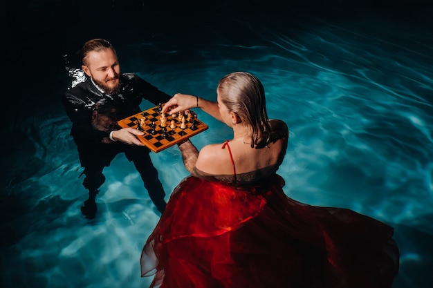 Un uomo vestito e una ragazza con un vestito rosso giocano a scacchi sull'acqua della piscina