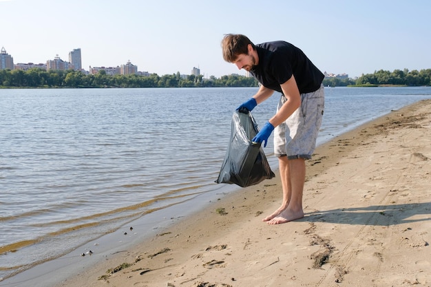 Un uomo volontario pulisce la spiaggia dalla spazzatura in una borsa nera. Pulizia delle spiagge della città