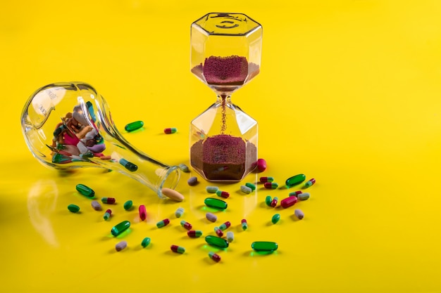 Un vaso trasparente con compresse che giace vicino a una manciata di compresse sparse di varie forme e colori accanto a una clessidra con grana rossa