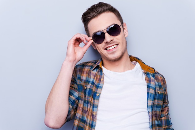 È un vero macho. Bel giovane in camicia che si aggiusta gli occhiali da sole e guarda la telecamera mentre si trova in piedi su uno sfondo grigio