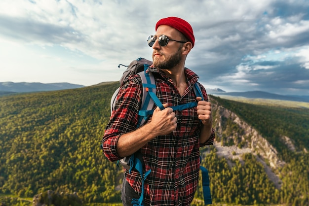 Un viaggiatore barbuto con uno zaino sulla cima di una montagna. Ritratto di un viaggiatore con un berretto rosso e occhiali da sole. Un turista con uno zaino si erge sullo sfondo di una montagna. Copia spazio