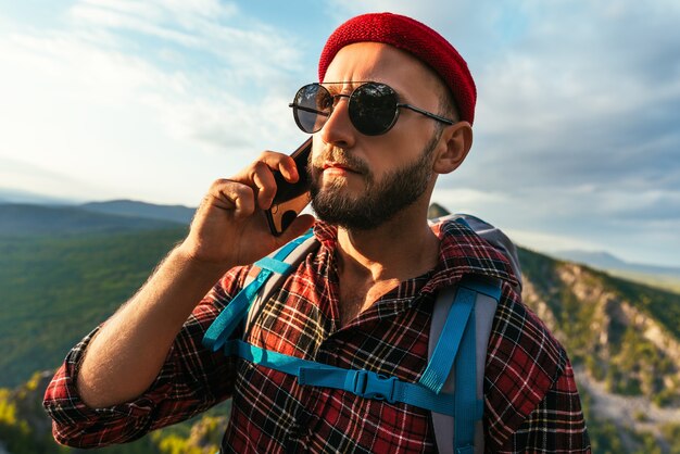 Un viaggiatore maschio con un cappello rosso con uno zaino sta parlando al telefono sullo sfondo delle montagne. Un uomo in abiti da trekking parla su uno smartphone al tramonto in montagna. Conversazione telefonica