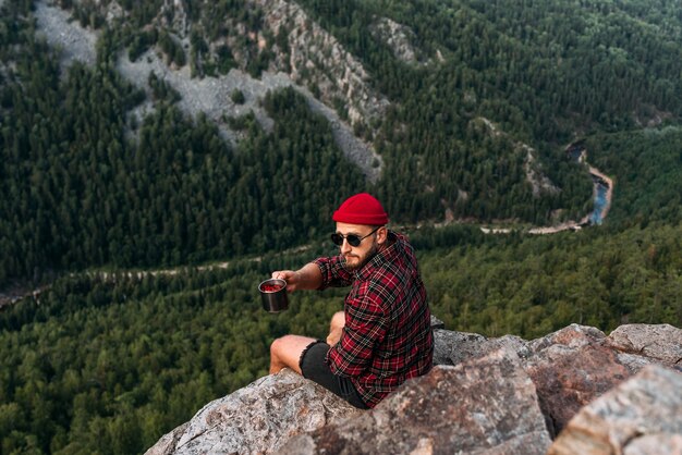 Un viaggiatore uomo seduto sul bordo di una scogliera. Un viaggiatore maschio è seduto su una roccia con una vista dall'alto della foresta. Il concetto di ricreazione e turismo. Un uomo gode di splendide viste. Viaggiare