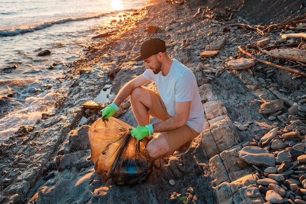 Un volontario di sesso maschile lega un sacco pieno di spazzatura sullo sfondo il mare Il concetto di conservazione ambientale e pulizia della zona costiera