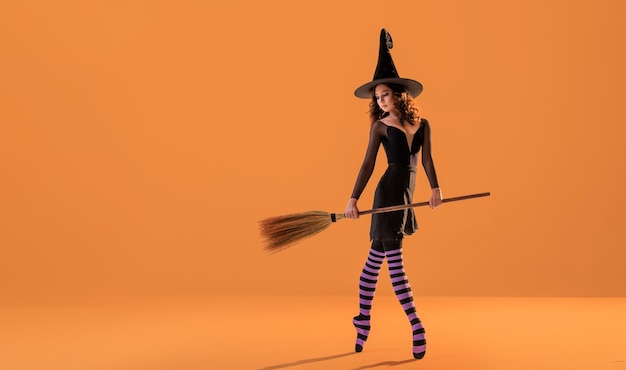Una ballerina su scarpe da punta in un costume da strega nera con un cappello e con una scopa balla su uno sfondo arancione