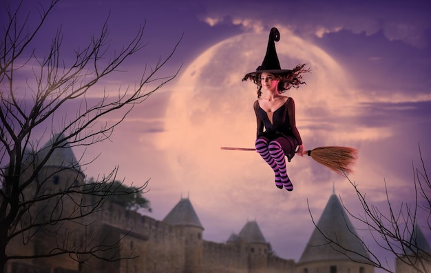 Una ballerina sulle scarpe da punta in un costume da strega nera con un cappello vola su una scopa sullo sfondo di un antico castello e un'enorme luna lilla Balletto di Halloween