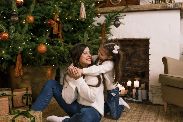 Una bambina abbraccia la sua amorevole madre seduta per terra sotto un albero di Natale. famiglia felice