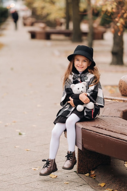 Una bambina alla moda in abiti autunnali è seduta su una panchina nella città autunnale