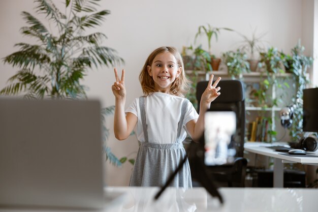 Una bambina allegra mostra gesti di pace in posa per la fotocamera del cellulare in camera