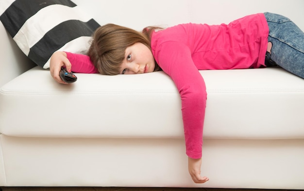 una bambina annoiata sdraiata sul divano guarda la TV durante la quarantena del Coronavirus