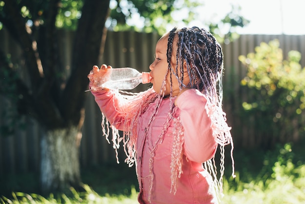 Una bambina bellissima con le trecce intrecciate beve acqua all'aperto in giardino