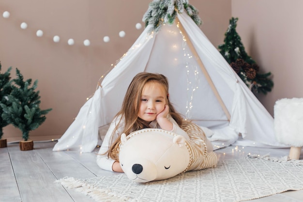Una bambina bellissima in pigiama bianco gioca con un orsacchiotto vicino alla stanza dei bambini del teepee con ca
