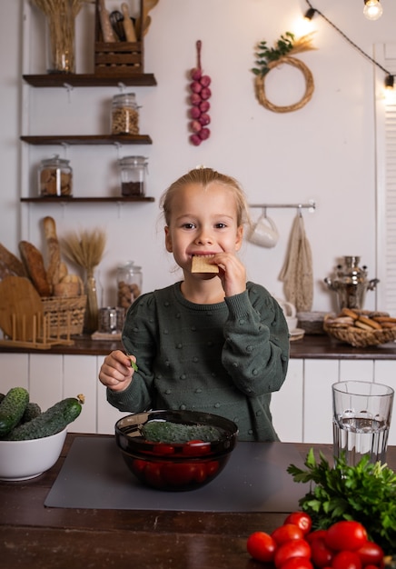 Una bambina bionda è seduta al tavolo della cucina con frutta e verdura e mangia pane