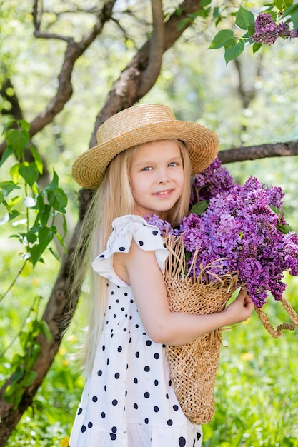 Una bambina bionda felice con un cappello di paglia tiene in mano un cesto di fiori lilla